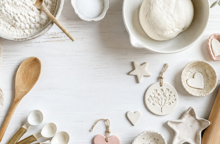 Baking ingredients, dough, and utensils are arranged on a white surface alongside handmade clay ornaments in heart and star shapes.